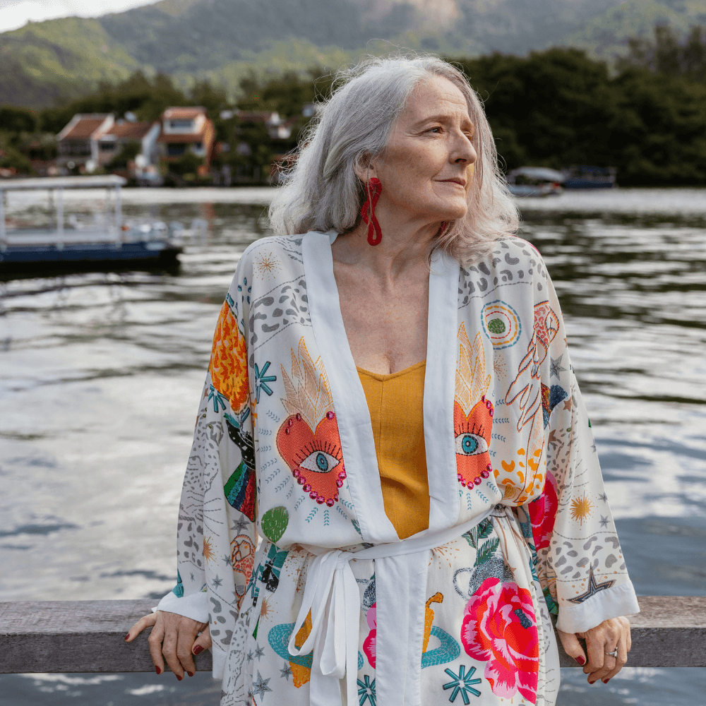 A grey-haired older woman in a patterned kimono resting on a wooden rail beside a calm harbour, looking out into the distance